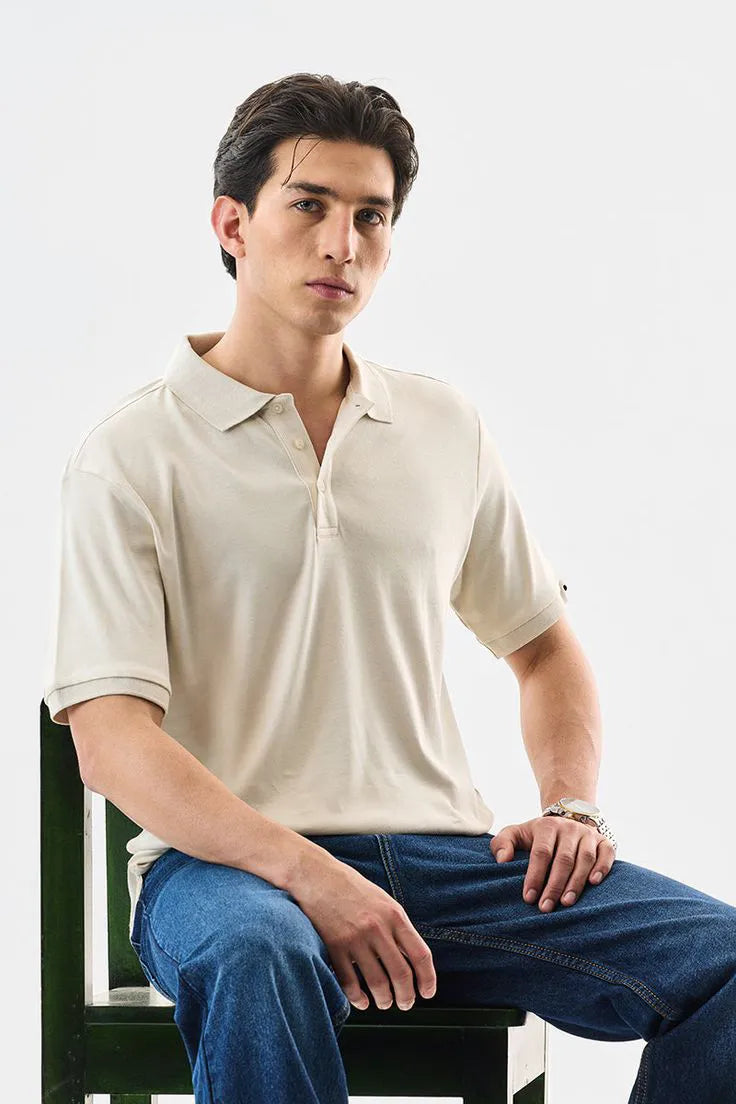 Man wearing a beige polo shirt and blue jeans sitting on a chair against a white background